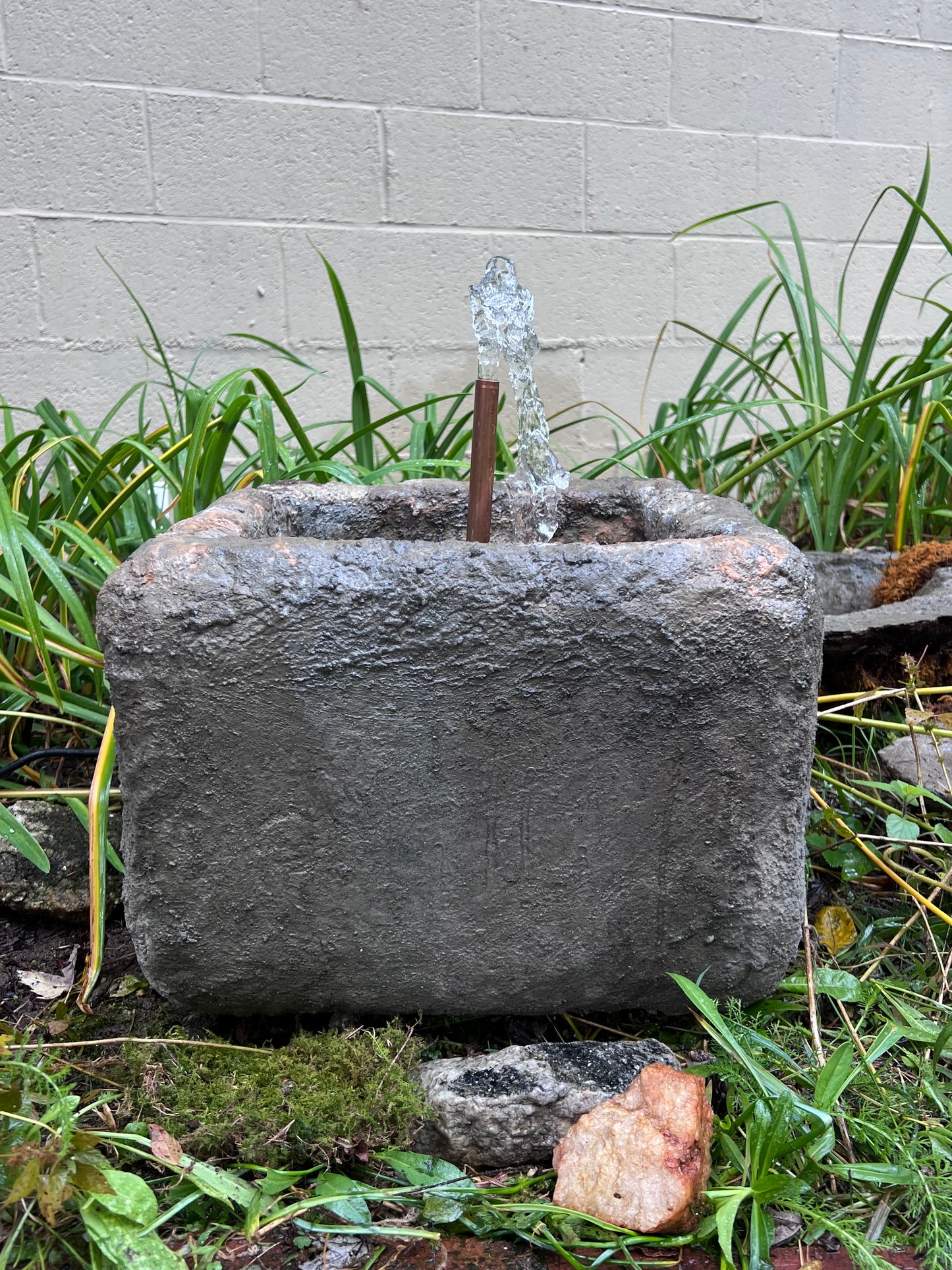 Decorative stone with water feature surrounded by grass and plants on a concrete surface