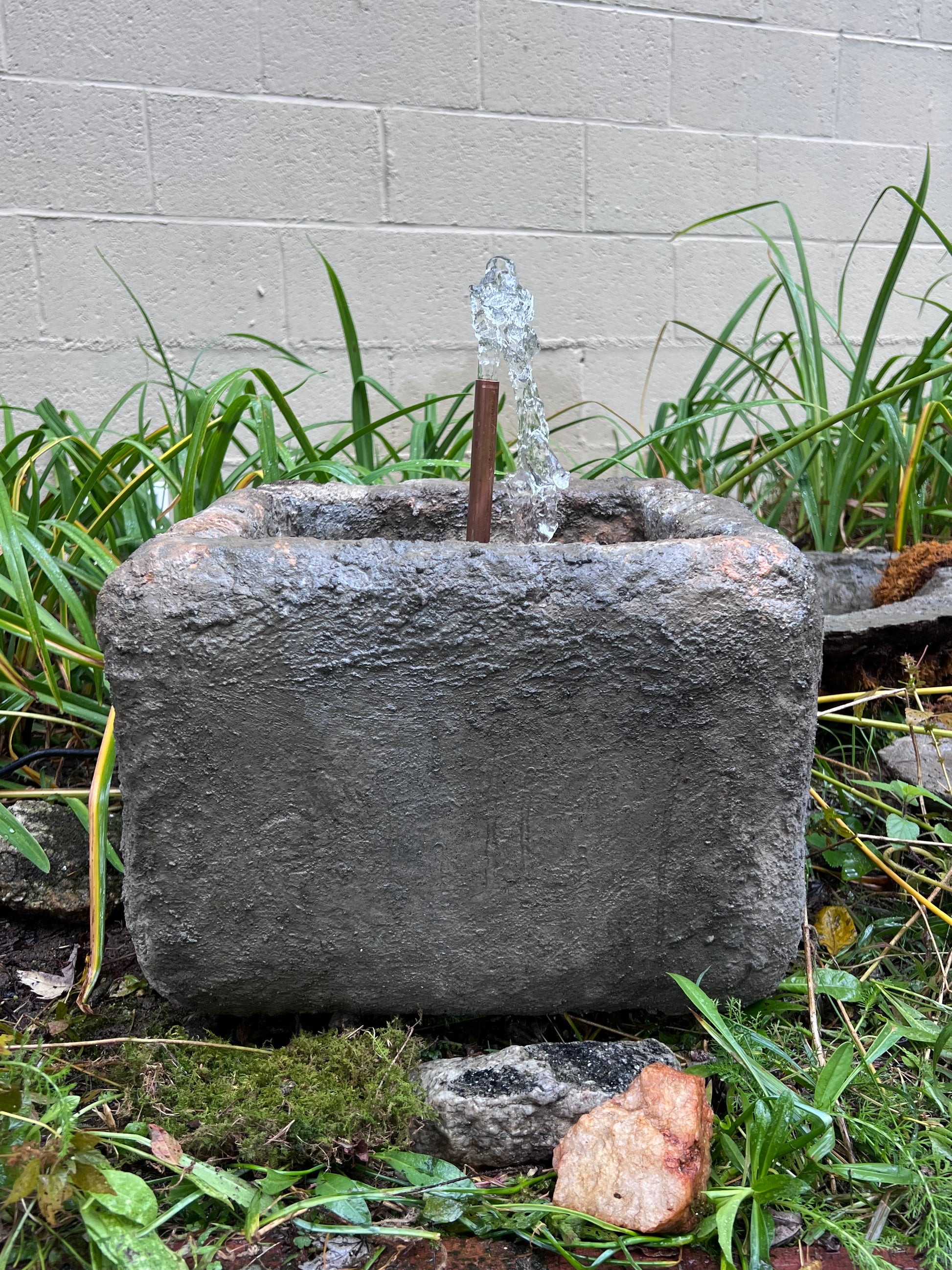 Decorative stone with water feature surrounded by grass and plants on a concrete surface