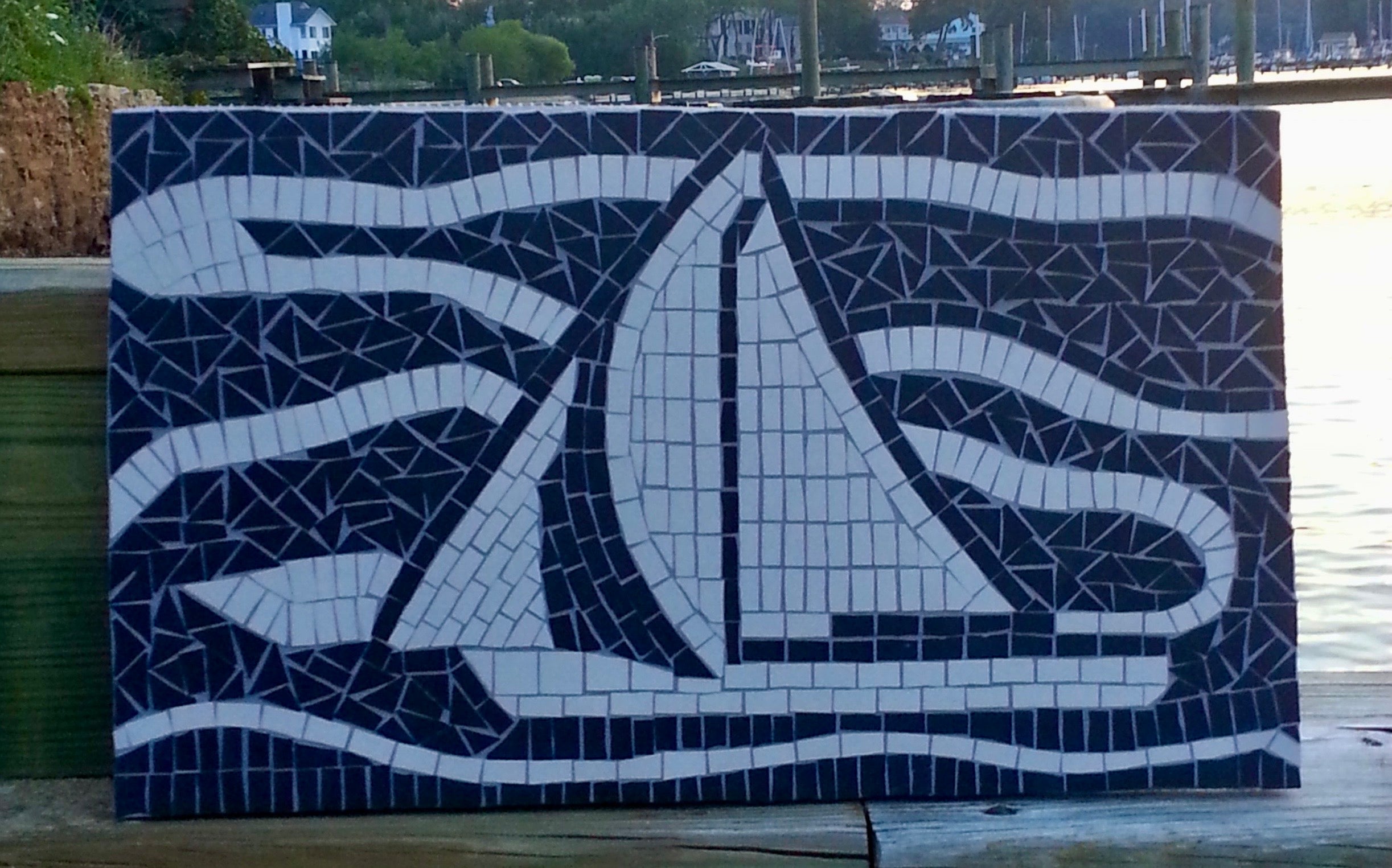 Mosaic artwork of a sailboat on a wooden surface with water and trees in the background
